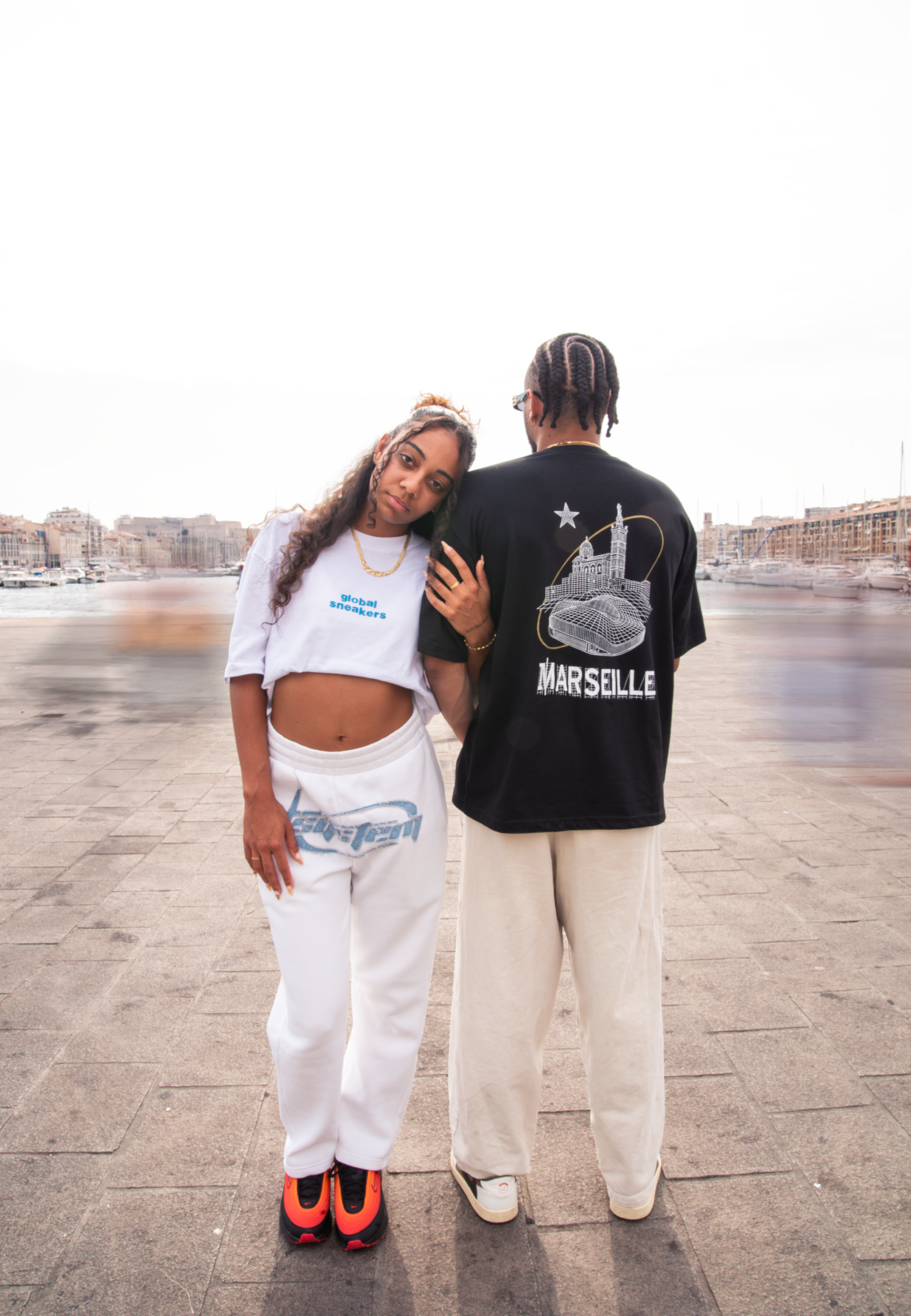 T-shirt noir Global Sneakers x Ailleurs/Higher Marseille – sérigraphie blanche et dorée avec monuments marseillais", porter par 2 mannequins tshirt noir et tshirt blanc et bleu