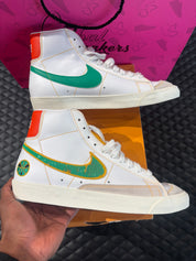 Nike Blazer Mid 77 Raygun d’occasion en état 9/10, cuir blanc, accents orange et jaune, silhouette montante classique, sneaker avec boîte.