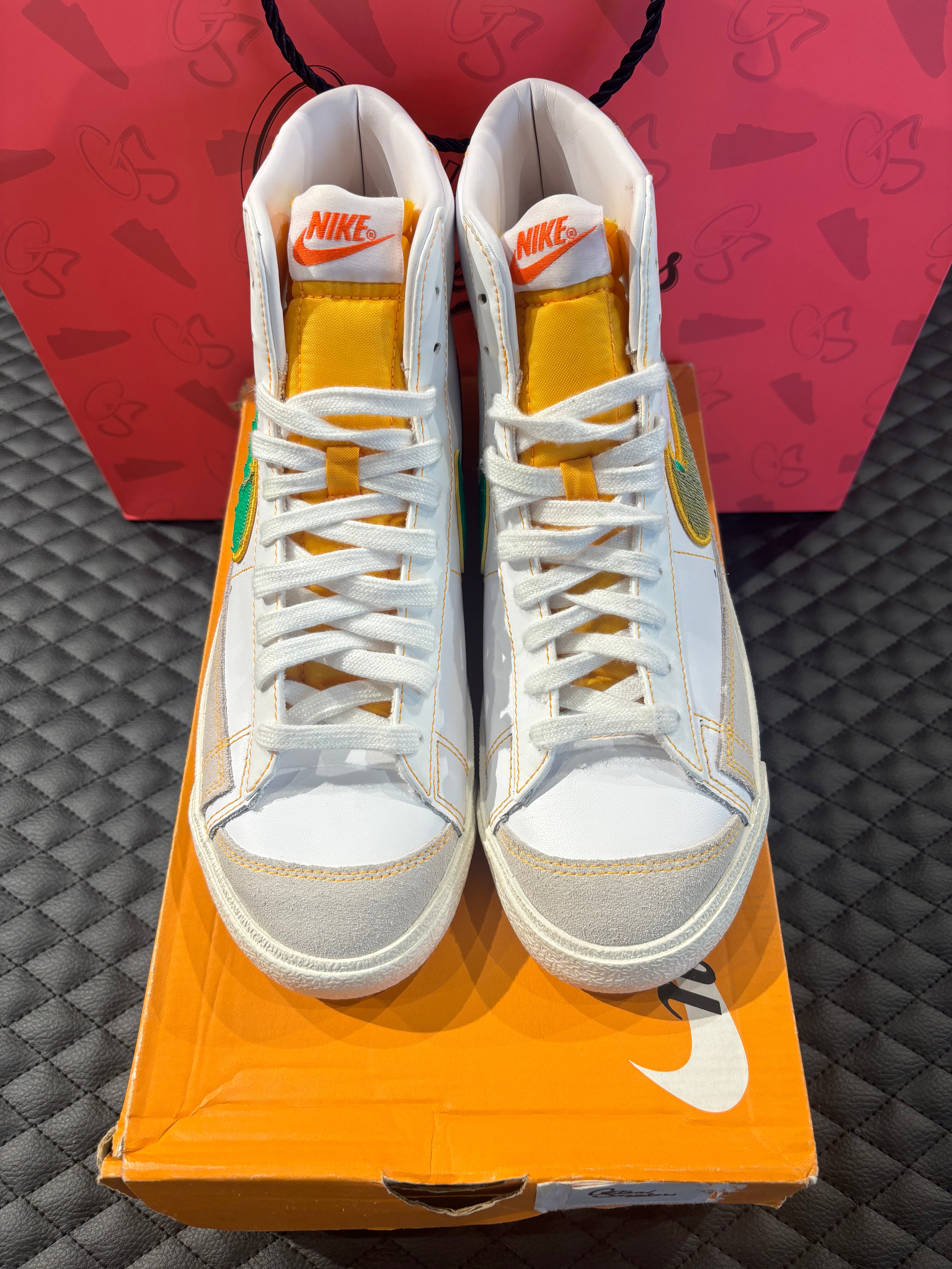 Nike Blazer Mid 77 Raygun d’occasion en état 9/10, cuir blanc, accents orange et jaune, silhouette montante classique, sneaker avec boîte.