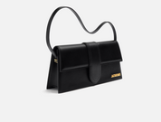 Jacquemus Le Bambino Long Shoulder Bag Black, sac d’épaule en cuir noir lisse avec rabat structuré, silhouette allongée et logo doré.