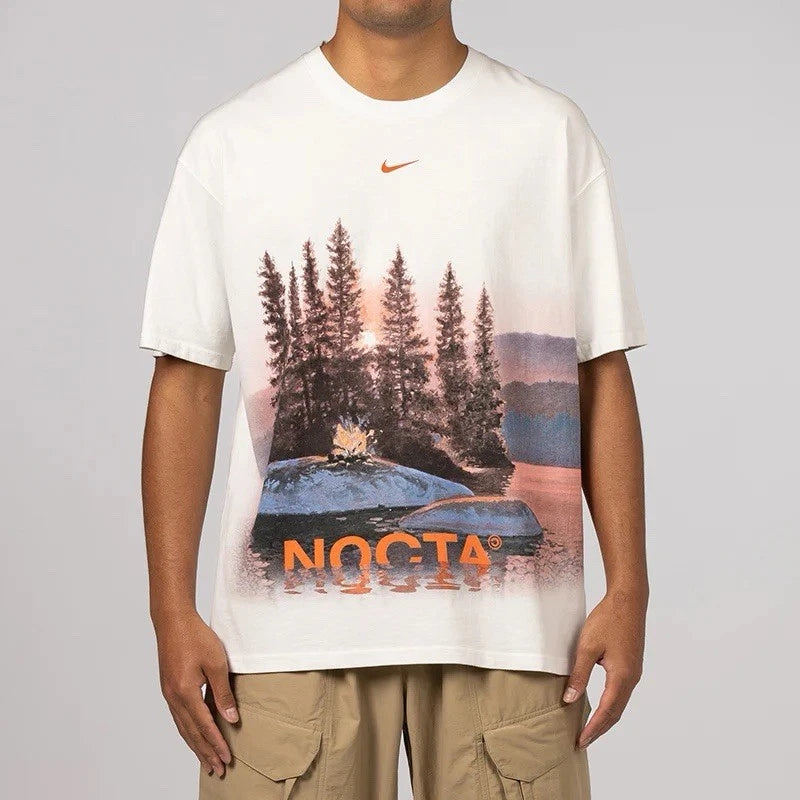 Nike x NOCTA Opal Reels Tee White, tee-shirt blanc en coton avec illustration paysage et pêcheur, détails multicolores et Swoosh orange.