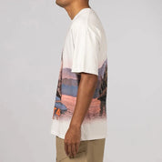 Nike x NOCTA Opal Reels Tee White, tee-shirt blanc en coton avec illustration paysage et pêcheur, détails multicolores et Swoosh orange.