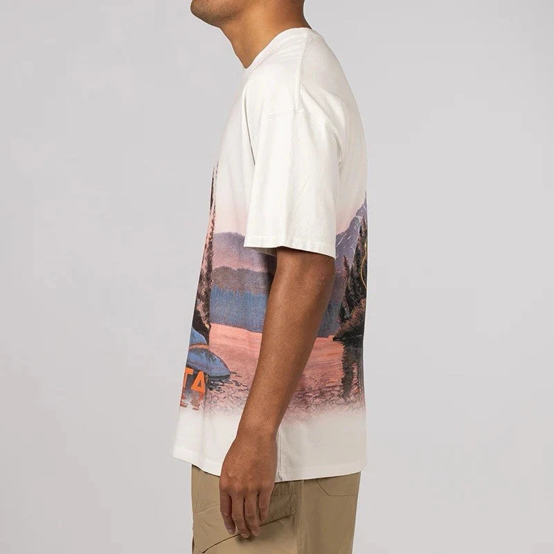 Nike x NOCTA Opal Reels Tee White, tee-shirt blanc en coton avec illustration paysage et pêcheur, détails multicolores et Swoosh orange.
