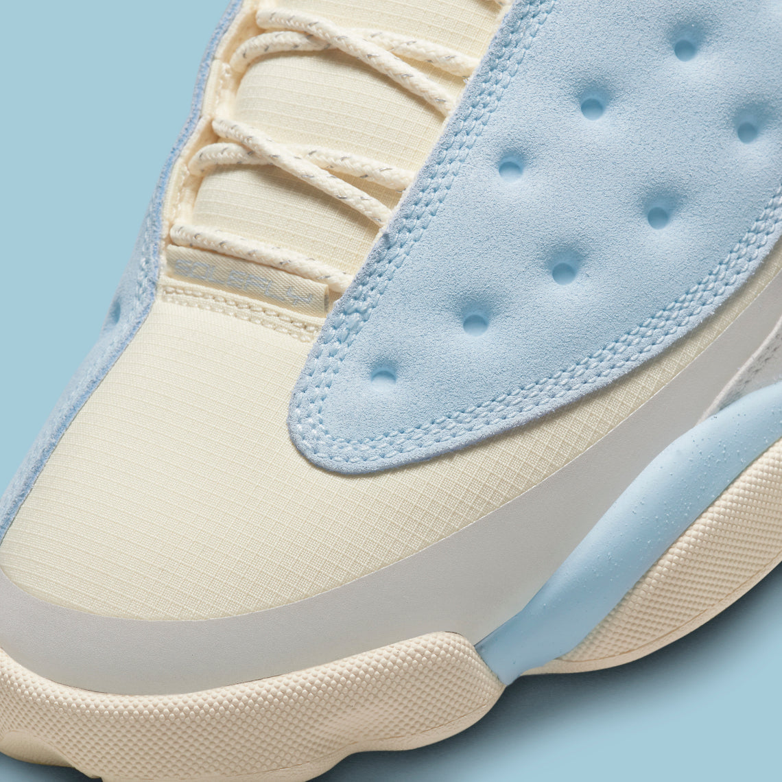 Vue zoomée sur la toe box de la Air Jordan 13 Retro SoleFly DX5763-100 en suède et ripstop blanc, gris et bleu ciel avec hologramme cat’s eye et semelle gum.