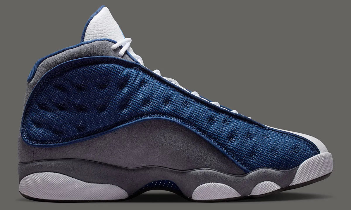 Air Jordan 13 Retro Flint 2026, sneaker Jordan Brand en cuir blanc, mesh réfléchissant bleu marine et suède Flint Grey avec amorti Zoom Air et œil holographique.