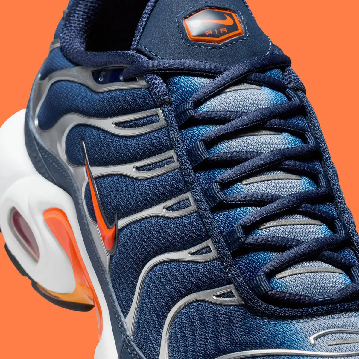 Vue zoomée sur les détails latéraux de la Nike Air Max Plus Syracuse bleu marine avec cage TPU gris argenté, Swoosh orange et unités Tuned Air visibles.
