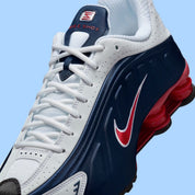 Nike Shox R4 USA 2025 avec tige en cuir synthétique blanc et bleu marine, détails Metallic Silver et Gym Red, amorti Nike Shox à quatre colonnes et semelle extérieure en caoutchouc.