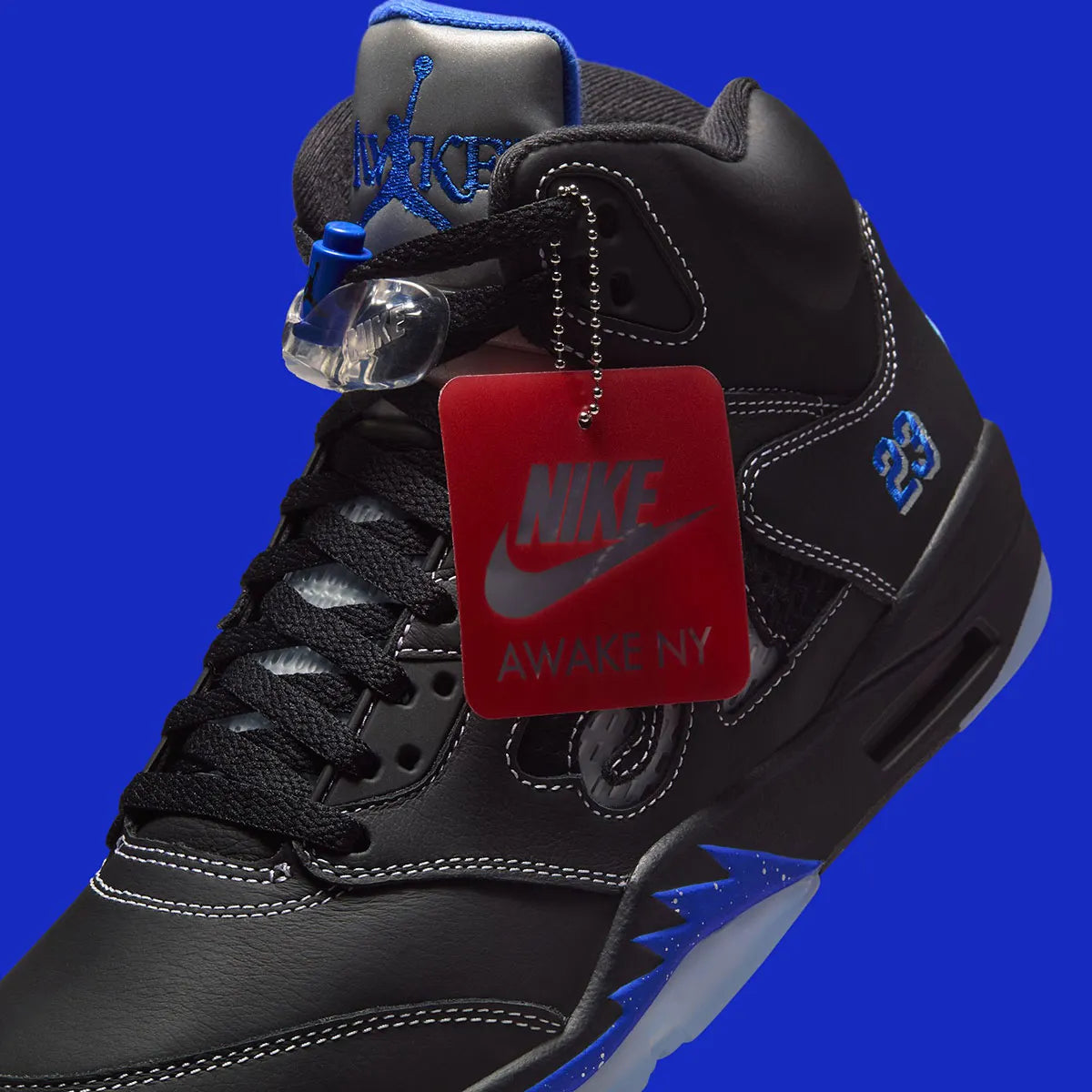 Vue latérale de la Air Jordan 5 Retro Awake NY Black DV4982-004, sneaker noire en cuir avec détails Racer Blue, languette argentée et logos Awake NY avec son étiquette. 