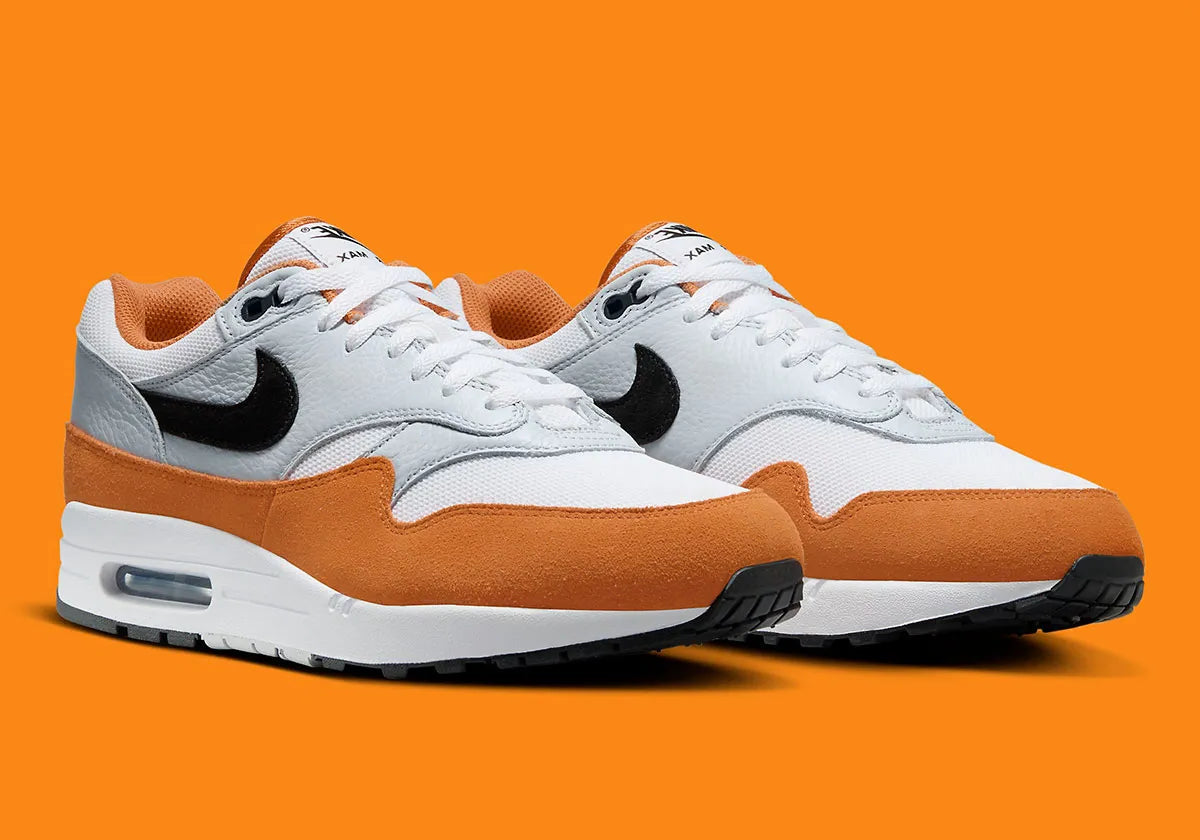 Nike Air Max 1 Monarch FN6952-101 en mesh blanc, cuir gris et suède orange Monarch avec Swoosh noir et unité Air visible.