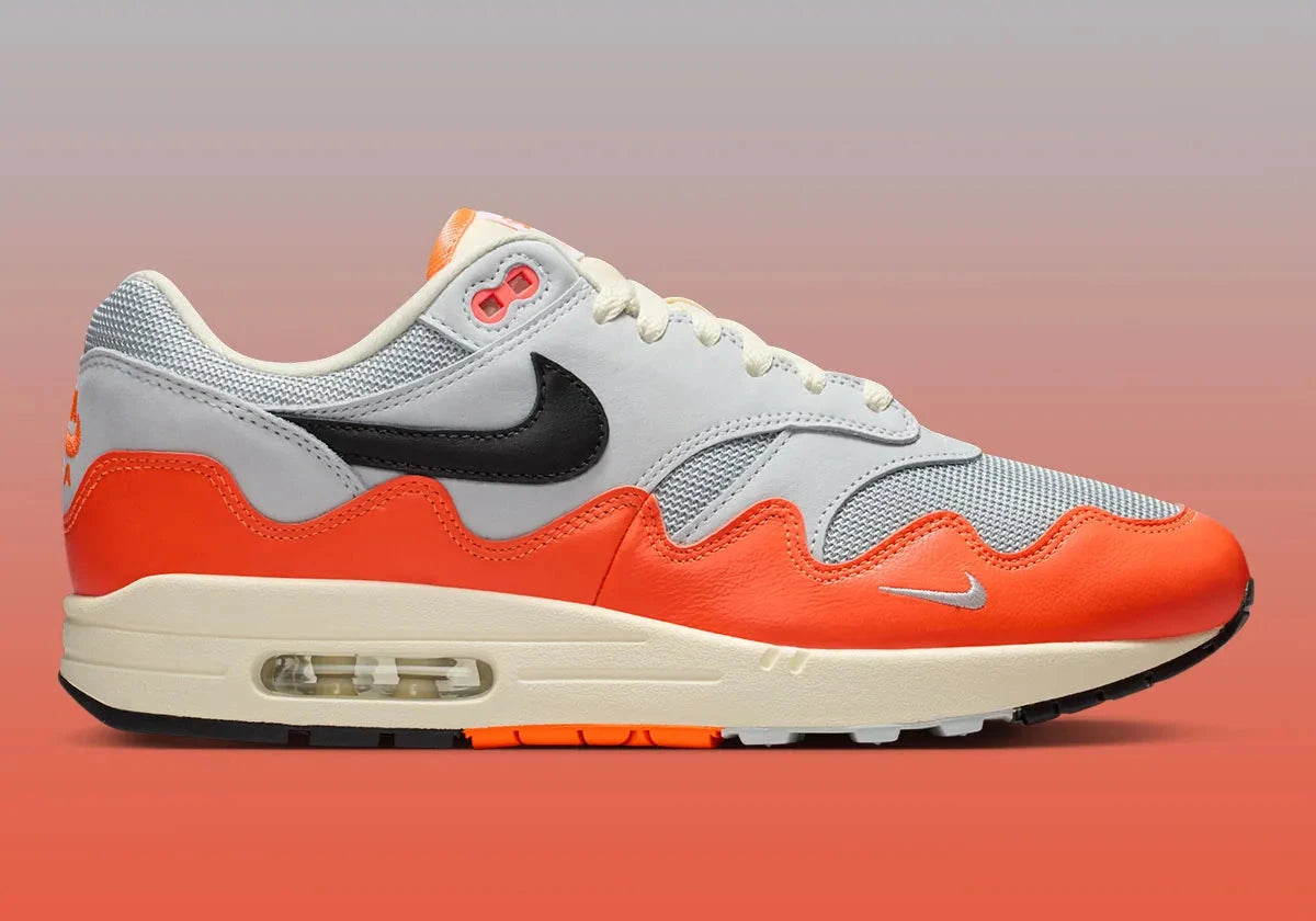 Sneakers Nike Air Max 1 Patta Waves Hyper Crimson Pure Platinum en mesh gris et cuir avec garde-boue ondulé orange et unité Air visible.