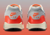 Sneakers Nike Air Max 1 Patta Waves Hyper Crimson Pure Platinum en mesh gris et cuir avec garde-boue ondulé orange et unité Air visible.