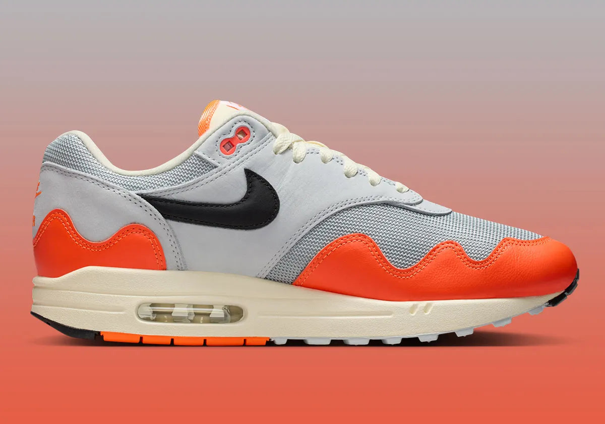 Sneakers Nike Air Max 1 Patta Waves Hyper Crimson Pure Platinum en mesh gris et cuir avec garde-boue ondulé orange et unité Air visible.