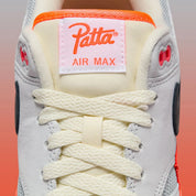 Sneakers Nike Air Max 1 Patta Waves Hyper Crimson Pure Platinum en mesh gris et cuir avec garde-boue ondulé orange et unité Air visible.